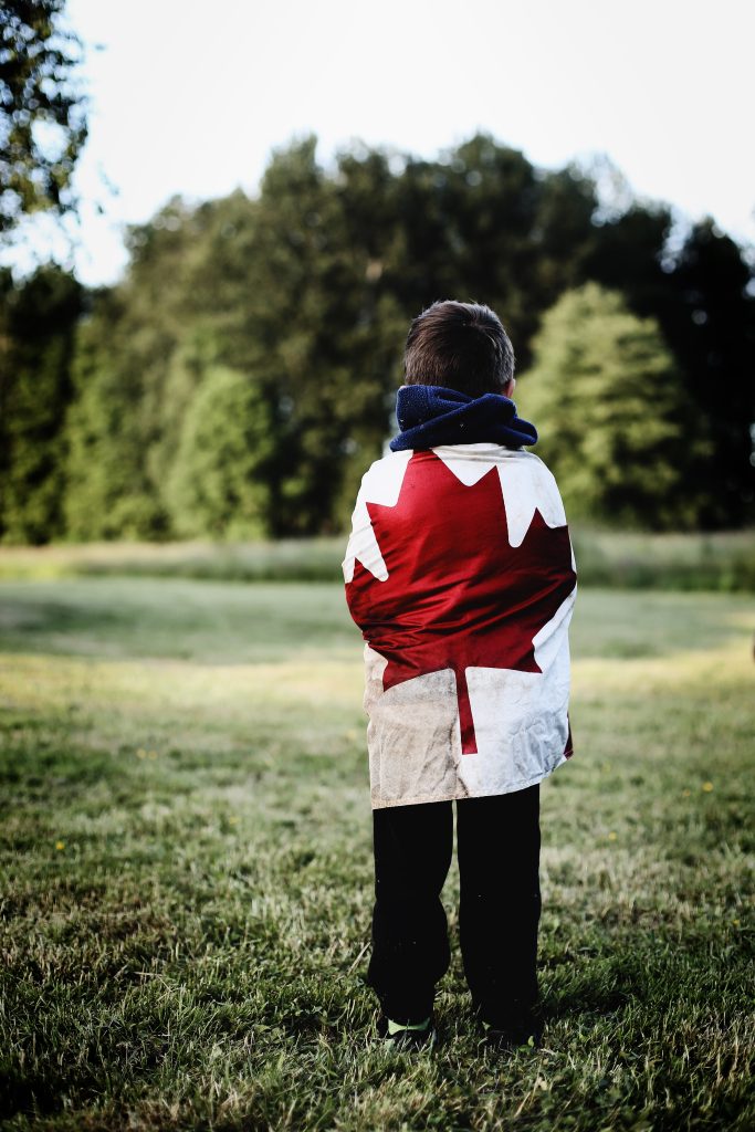 A young boy wrapped in a Canadian Flag looking out on a lake.