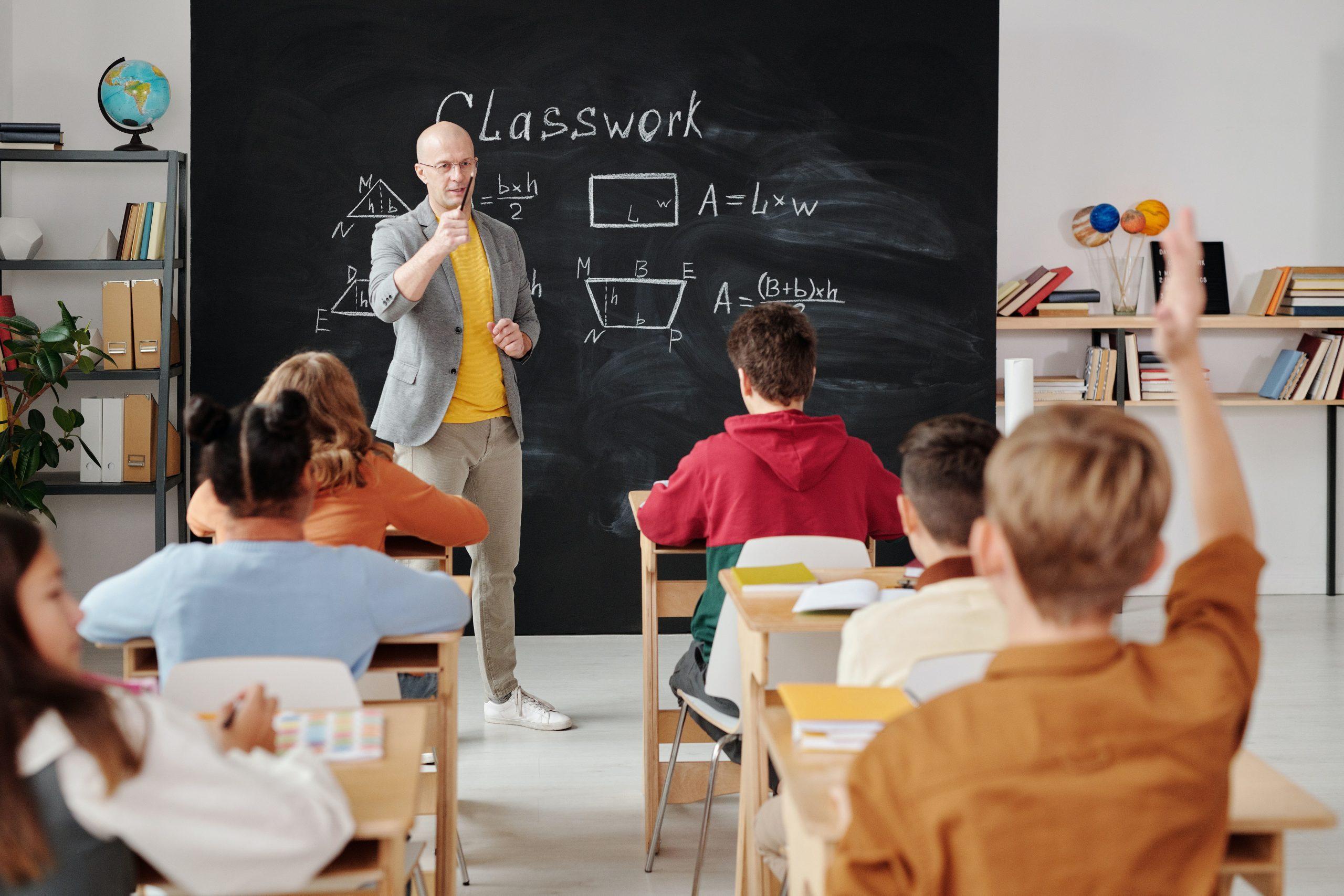 A teacher calls upon a young boy in an orange shirt with his hand raised.