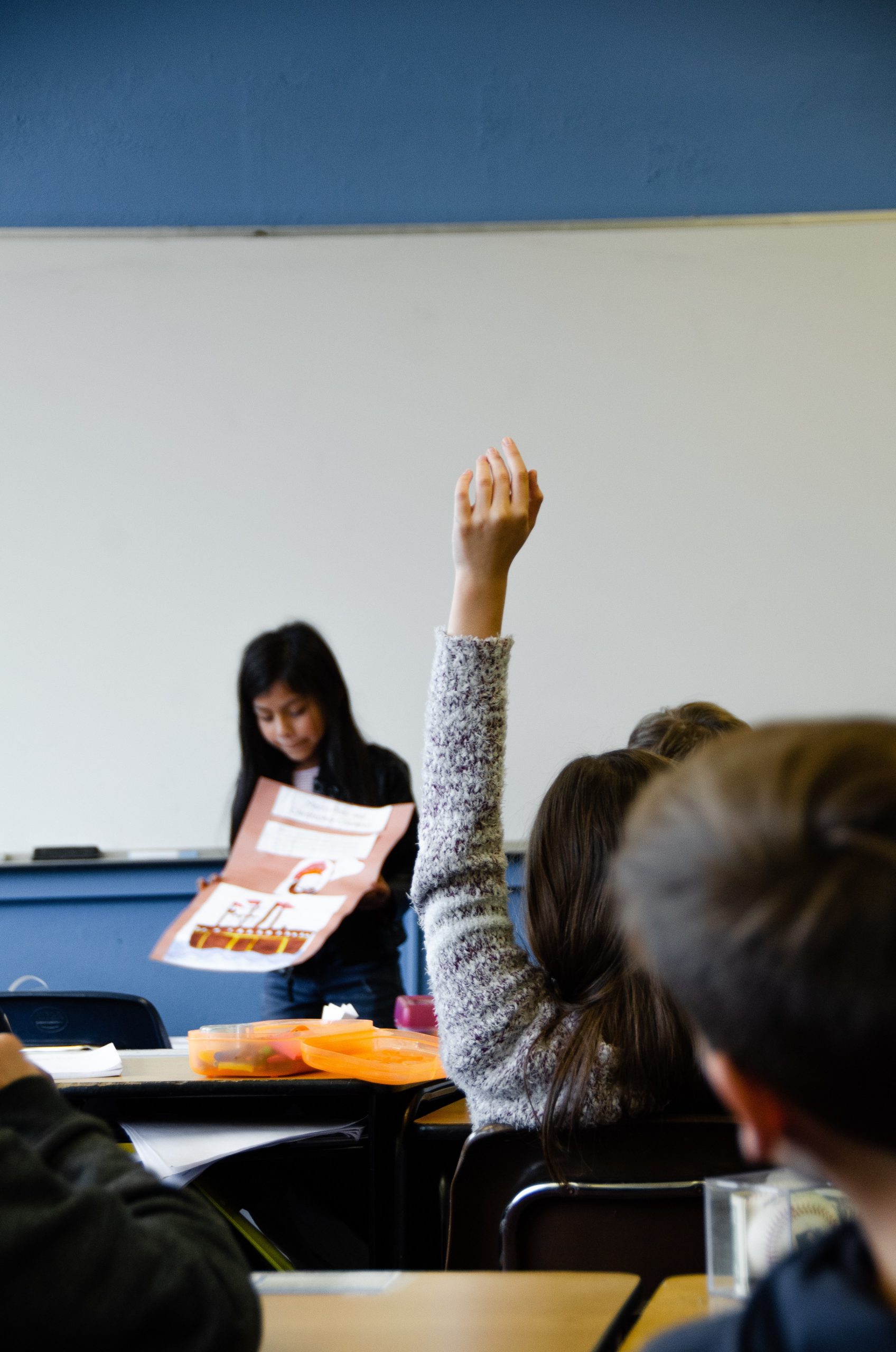 Student in grey sweater raising her hand in a classroom during a class presentation