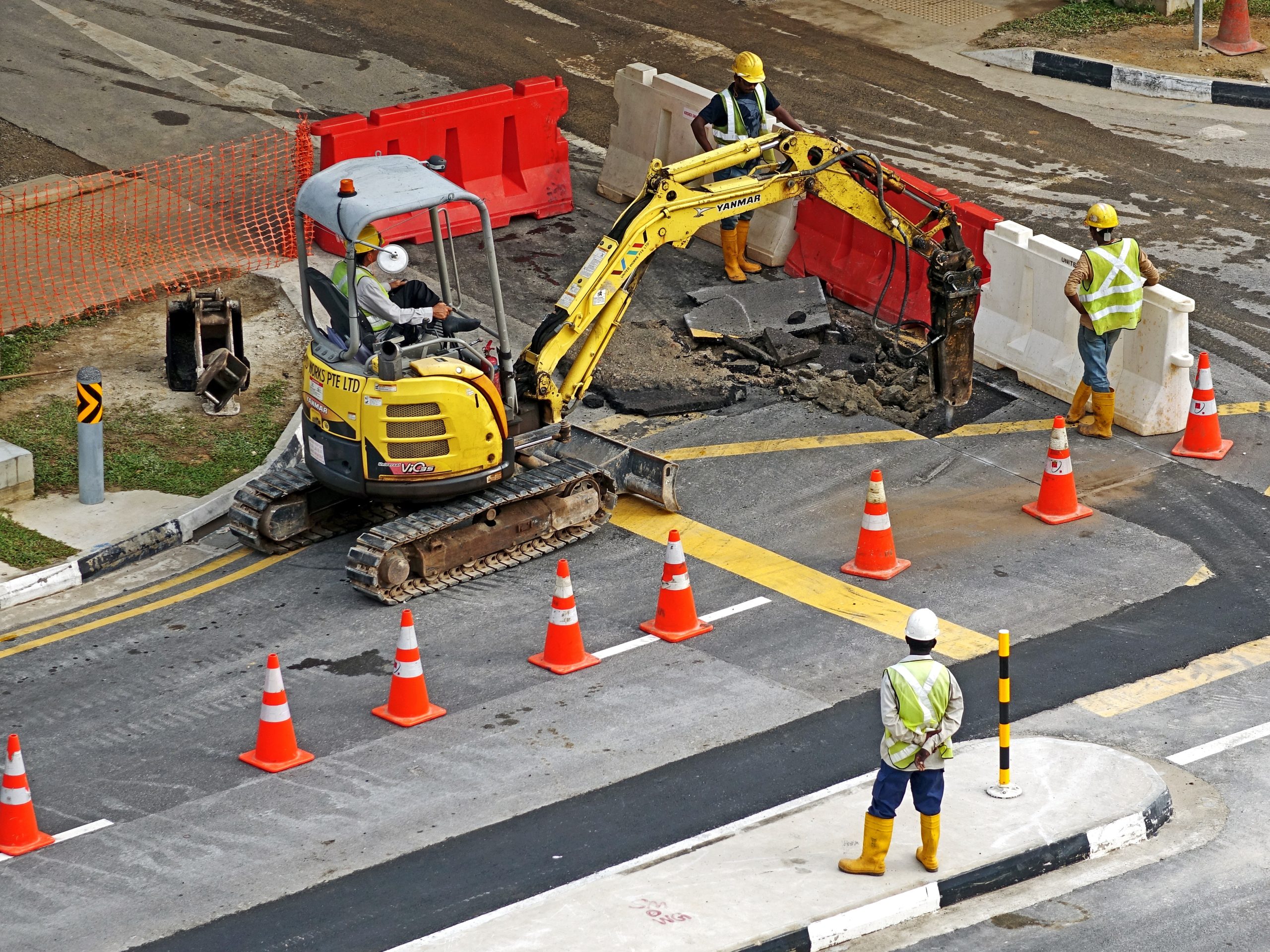 Excavator digging up a road while workers look on.
